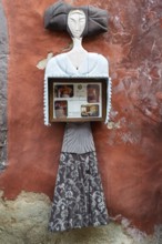 Abstract sculpture of a woman on a rust-red plastered wall. Figure holding a box with photos,