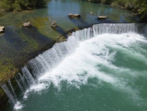 A wide waterfall with clear turquoise water, surrounded by rocks and green vegetation, aerial view,