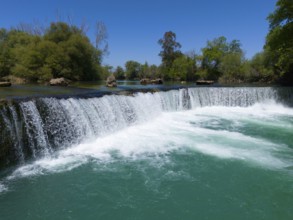 A waterfall flows into a turquoise-coloured river, surrounded by green vegetation and blue sky,