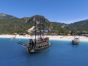 Pirate ship sails in front of a picturesque coastal landscape with mountains and clear blue water,