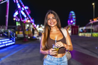 Young woman holding a refreshing drink at a lively amusement park, enjoying the vibrant atmosphere