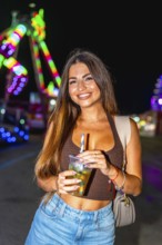 Young woman holding a refreshing cocktail at an amusement park, enjoying the vibrant night