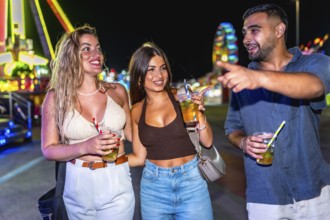 Group of friends enjoying cocktails, walking and laughing together at an amusement park, surrounded