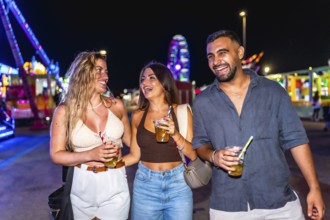 Three friends walking and laughing together at amusement park at night, holding drinks and enjoying