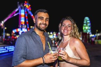 Young man and woman smiling, holding cocktails, enjoying a night out at amusement park with