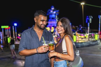 Young couple smiling and holding colorful cocktails at a lively funfair, celebrating nightlife and