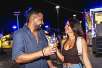 Young man and woman smiling and toasting with colorful cocktails while enjoying the vibrant