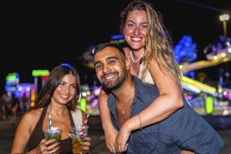 Three friends are having fun at an amusement park at night, enjoying cocktails and the festive