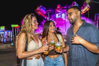 Three friends are holding cocktails and chatting at an amusement park at night, enjoying the