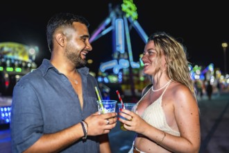 Happy couple enjoying drinks at amusement park, celebrating summer night with colorful lights and