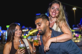 Three friends are having cocktails at an amusement park at night, enjoying the vibrant atmosphere