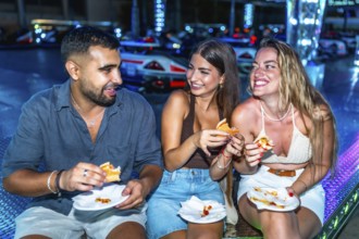 Three happy friends are enjoying burgers and pizza at an amusement park, illuminated by colorful