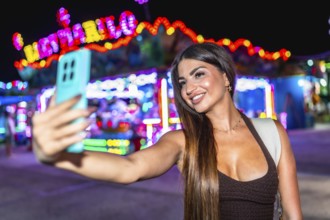 Happy woman enjoying a vibrant night at an amusement park, taking a selfie with her smartphone