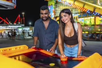 Happy young couple enjoying a lively night out, playing air hockey together at a colorful amusement