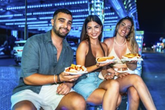 Three friends are sitting and smiling, holding hot dogs on paper plates at a brightly lit amusement