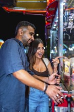 Young couple having fun playing a claw crane game at an amusement park at night, trying to win a
