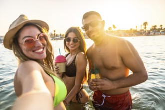 Group of cheerful young friends taking selfie in the sea while drinking cocktails at sunset during