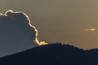A radiant sunset conjures up warm colours as the clouds hang over a mountain range. Trees line the