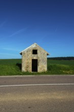 A rustic stone hut stands alone by a quiet road, surrounded by vibrant green fields. Auvergne.