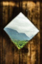 Rustic wooden frame reveals a lush green landscape with mountains in the background under a cloudy