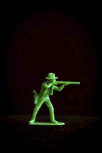 A vibrant green plastic toy cowboy stands in a tactical pose, aiming a rifle against a dark