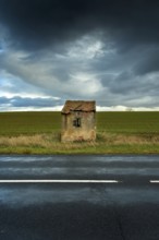 A small, dilapidated hut with a broken roof and cracked walls is positioned next to a quiet road.