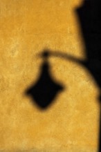 Soft afternoon light creates a striking shadow of a lantern on a textured yellow wall. The warm