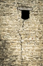 A weathered stone wall displays a prominent crack running vertically, featuring a small window.