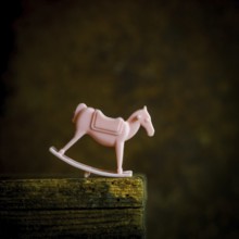 A small pink rocking horse toy sits on a weathered wooden shelf, gently illuminated in low light.