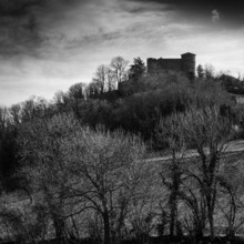 A historic castle stands prominently on a hill, silhouetted against a moody sky at dusk. Bare trees