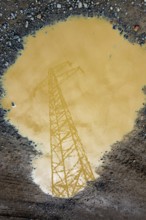 A power line tower is reflected in a large puddle of muddy water. The cloudy sky is visible, adding