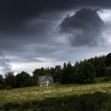 A solitary house stands in a lush green field under a sky filled with dark, ominous clouds.