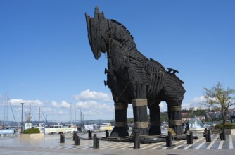 Large black wooden statue of a horse at the harbour with ships and blue sky in the background,