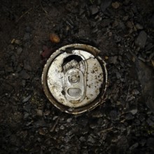 An aluminum can lies abandoned on dark, rich soil interspersed with small pebbles, underscoring the