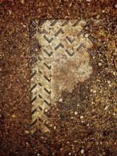A worn metal cover is partially submerged in dirt and gravel, showcasing intricate patterns from