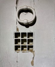 A striking white door knocker adorns a weathered wall featuring small square openings arranged in