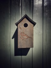 A wooden birdhouse, featuring a carved bird design, is suspended from a rustic gray wall. This