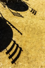 Shadows cast by chairs and tables stretch across a sunlit ground, revealing intricate patterns on