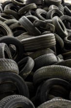 A large collection of worn tires is heaped together in a cluttered industrial setting. The scene
