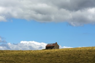 A stone cottage rests on a grassy slope, surrounded by expansive hills. The sky features vibrant