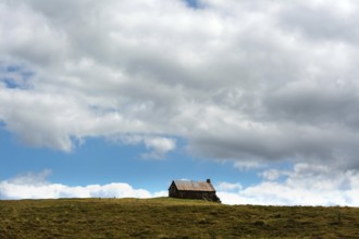 A small cabin sits alone atop a grassy hill, surrounded by wide-open space. The sky is filled with