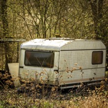 A weathered caravan stands abandoned in a quiet field, partially hidden by tall grass and wild
