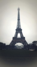 Eiffel Tower stands tall against a grey sky in Paris, surrounded by landscaped gardens. Visitors