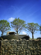 A vintage convertible car sits next to a stone wall, surrounded by lush green trees. The bright