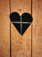 A rustic wooden door features a heart-shaped cutout that reveals iron bars behind it. The warm