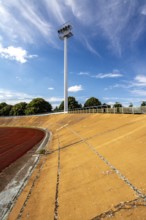 Bright daylight reveals an empty sports stadium with a vibrant red track. The tall floodlights,