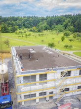 Partly completed building with workers in a landscaped environment, surrounded by forest, green