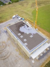 Building in shell construction with crane and vehicles on an open plot with surrounding fields,