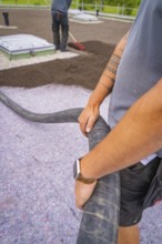 Worker holds a hose with visible tattoos and uses construction clothing, roof greening, house