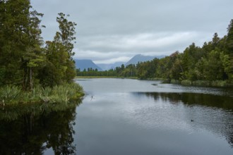 A peaceful lake surrounded by trees, with mountains on the horizon under a cloudy sky, summer, Lake